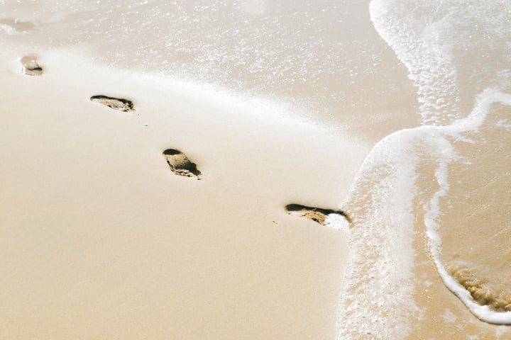 Footsteps in the sand being covered by a wave. 