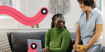 Two smiling women sitting around a laptop drinking coffee.